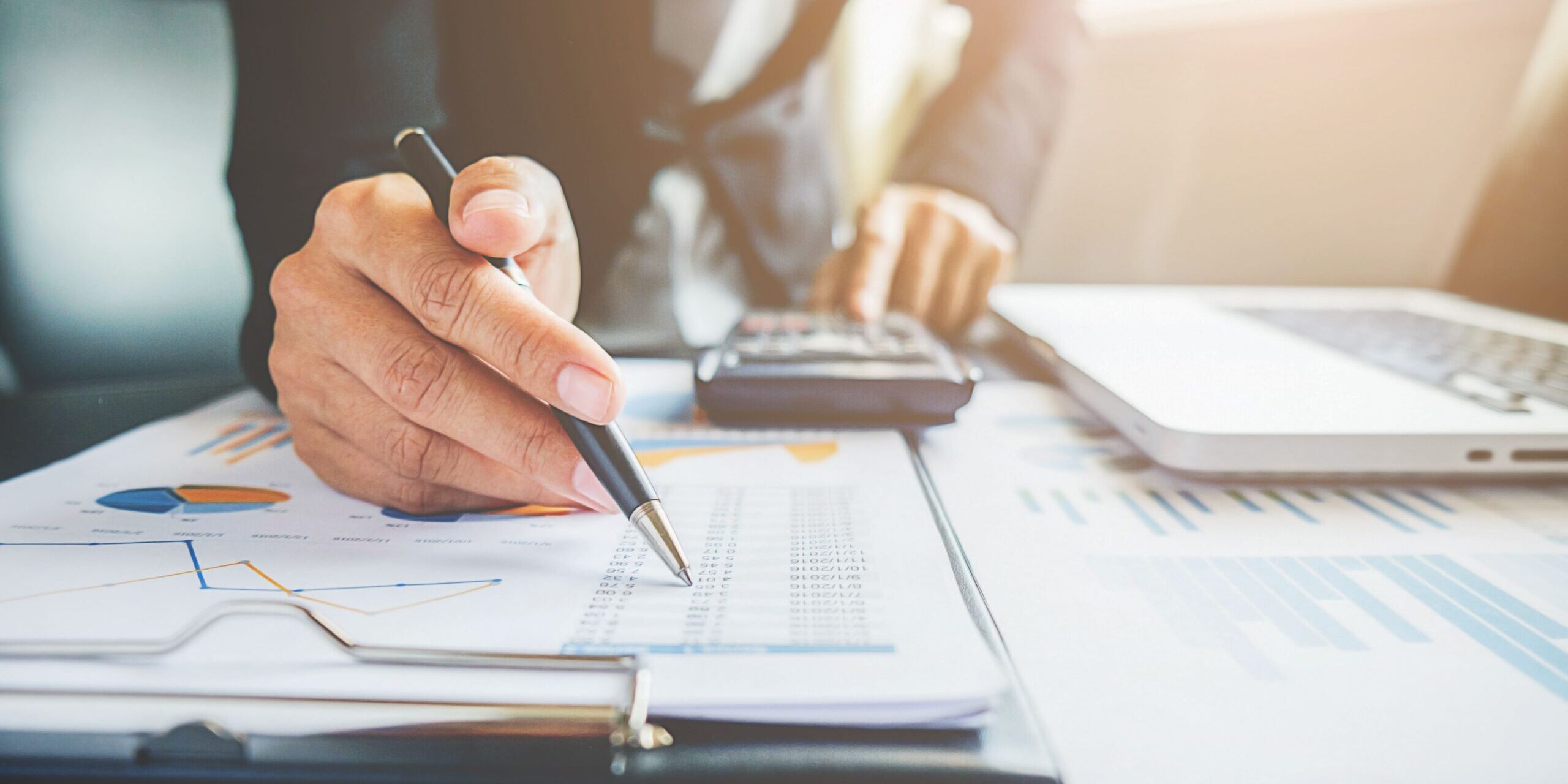 mesa com papéis, um notebook, calculadora e mãos de um homem. uma das mãos está com uma caneta direcionada ao papel e a outra digitando na calculadora para representar empréstimo para empresas